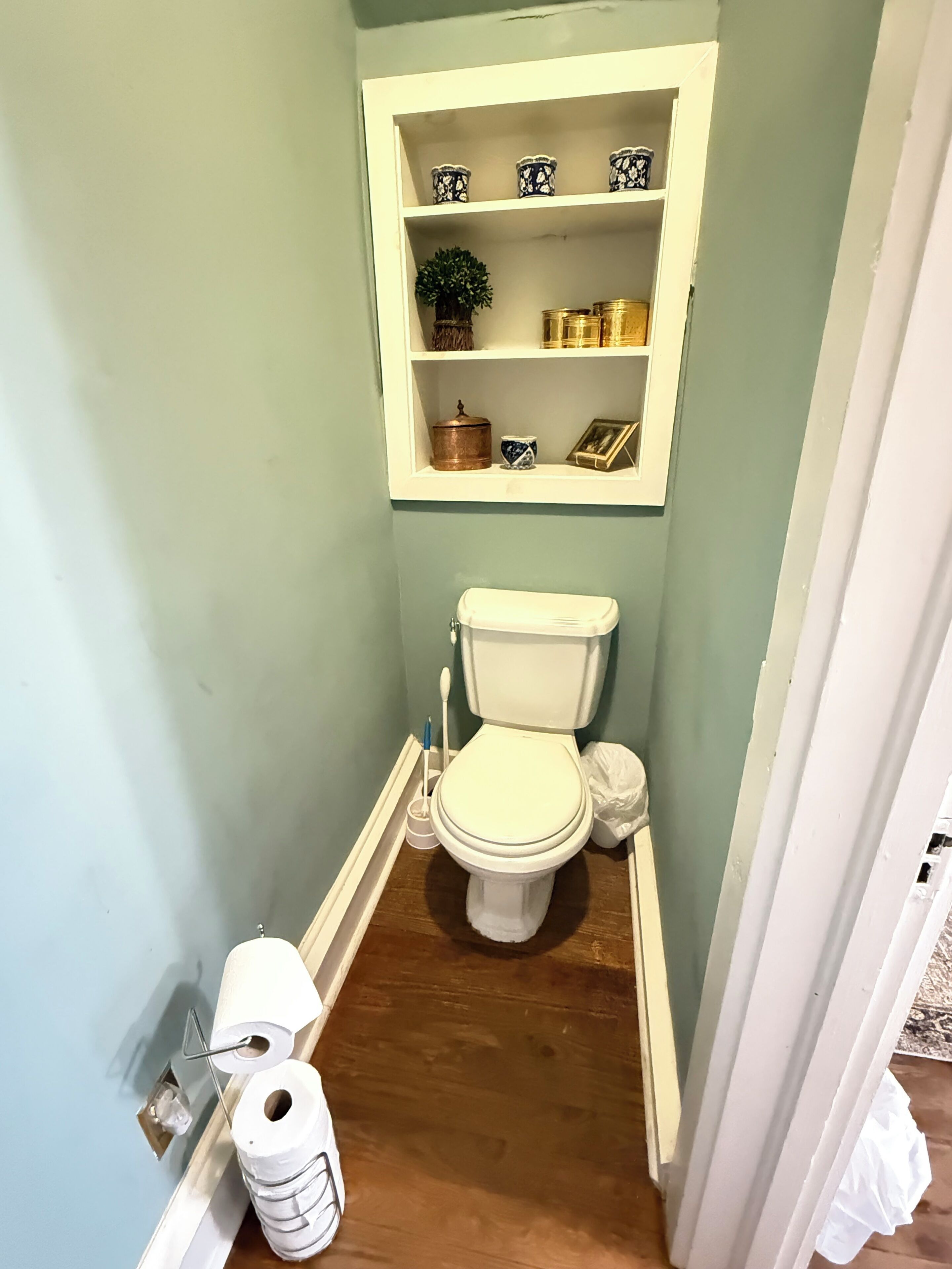 A small bathroom with a toilet, a shelf above it containing decorative items, and a roll of toilet paper on the floor.