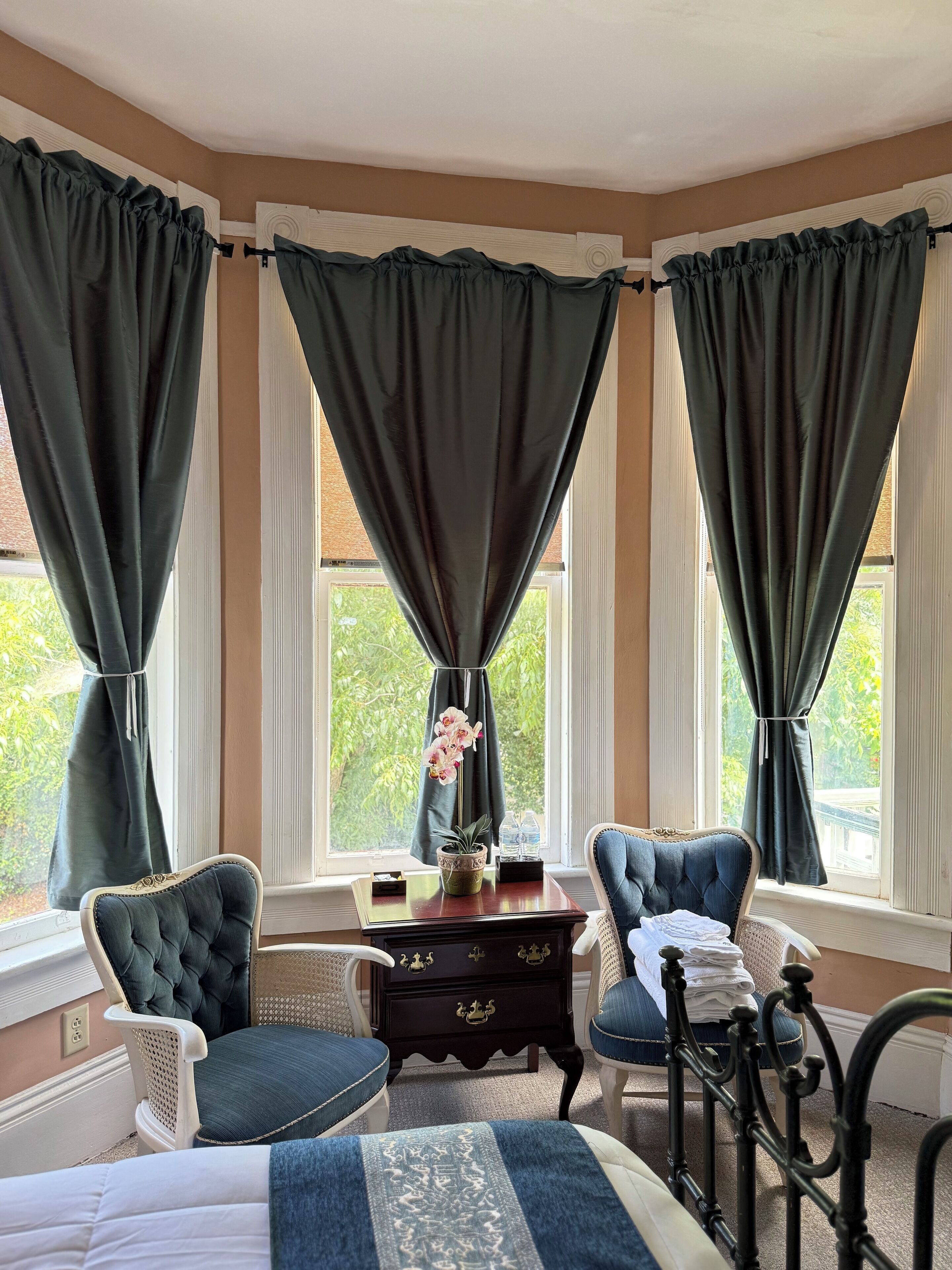 A room with a bed, two chairs, a small table with a vase of flowers, and a view of greenery outside.