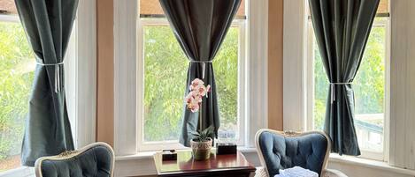 A room with a bed, two chairs, a small table with a vase of flowers, and a view of greenery outside.