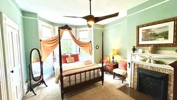 Una cama con dosel, un ventilador de techo, una chimenea, un espejo y una ventana con cortinas drapeadas.