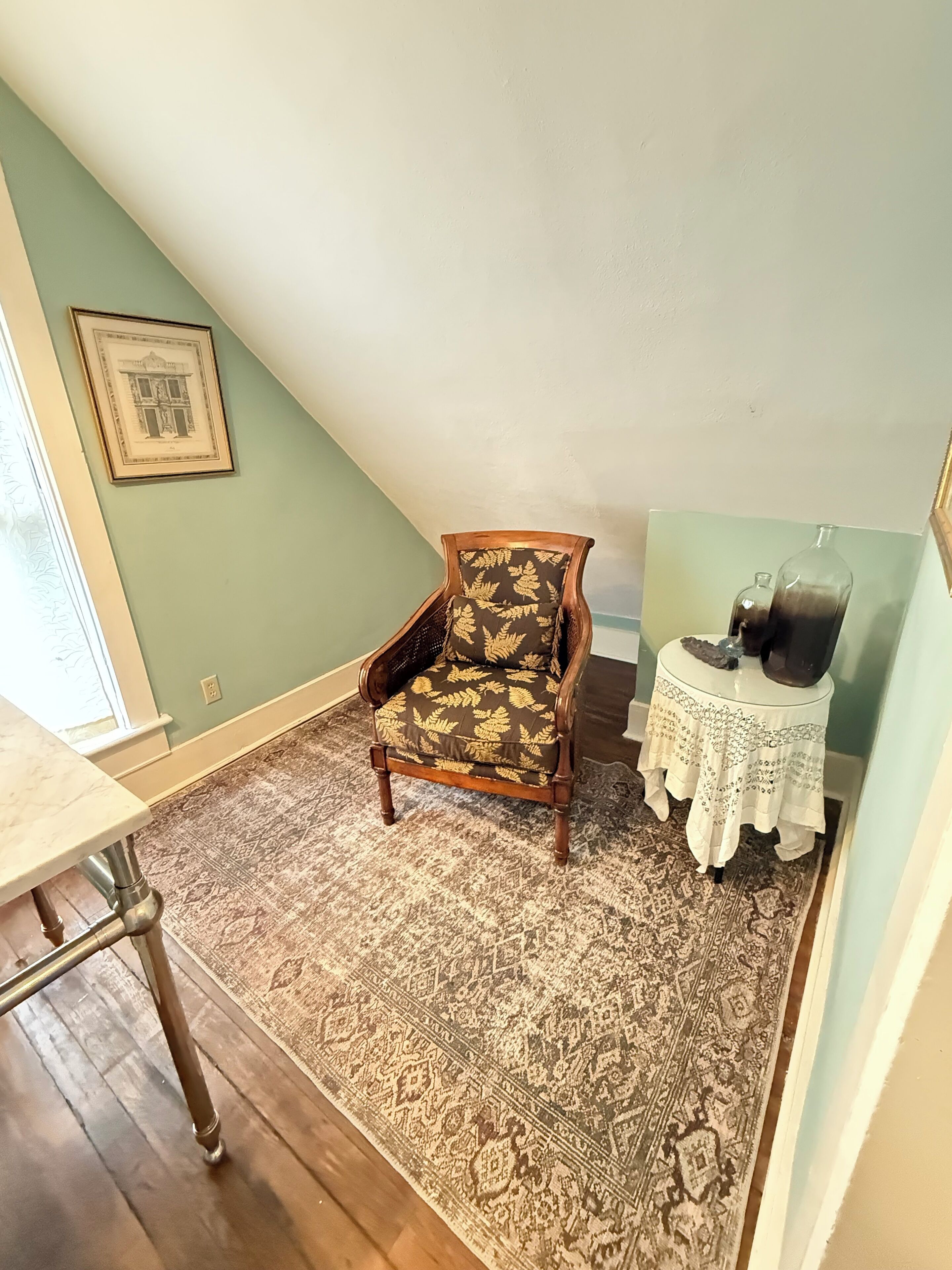 A room with a patterned rug, a wooden chair, a small table with a glass bottle, and a framed picture on the wall.