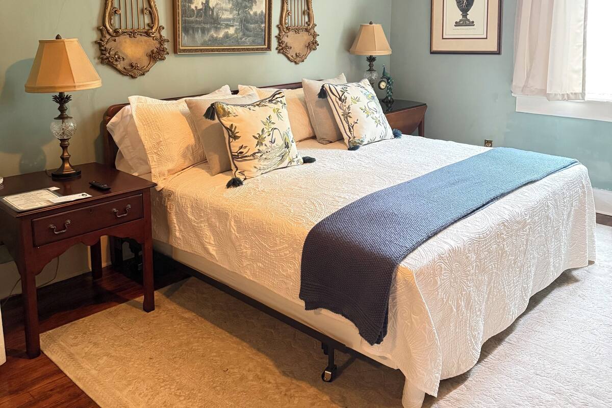 Quarto com cama, mesas de cabeceira, abajur, quadro emoldurado e janela com cortinas.