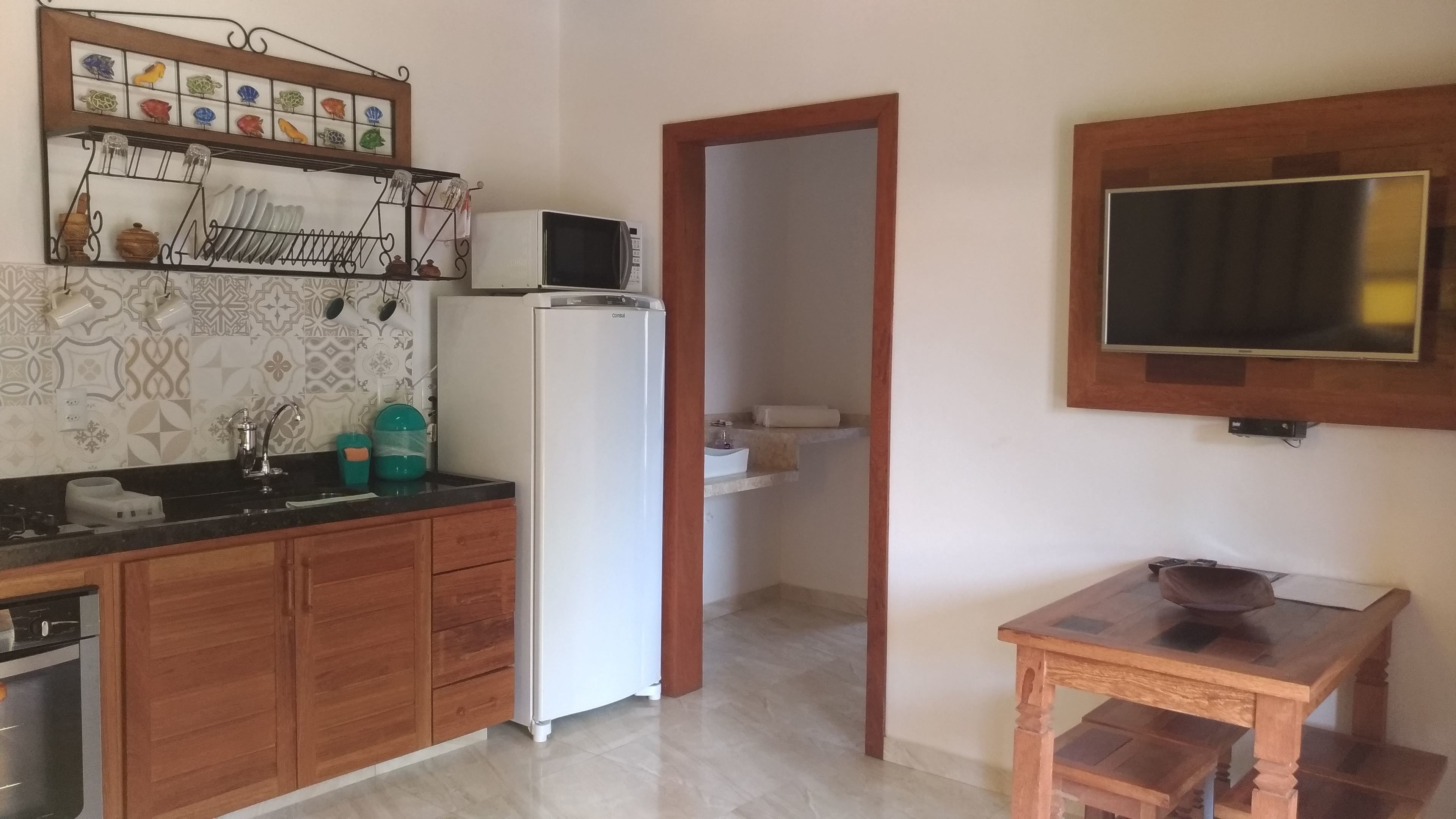 A kitchen with wooden cabinets, a white refrigerator, a microwave, and a sink. There is a small wooden table with a bowl. A TV is mounted on the wall above the table.