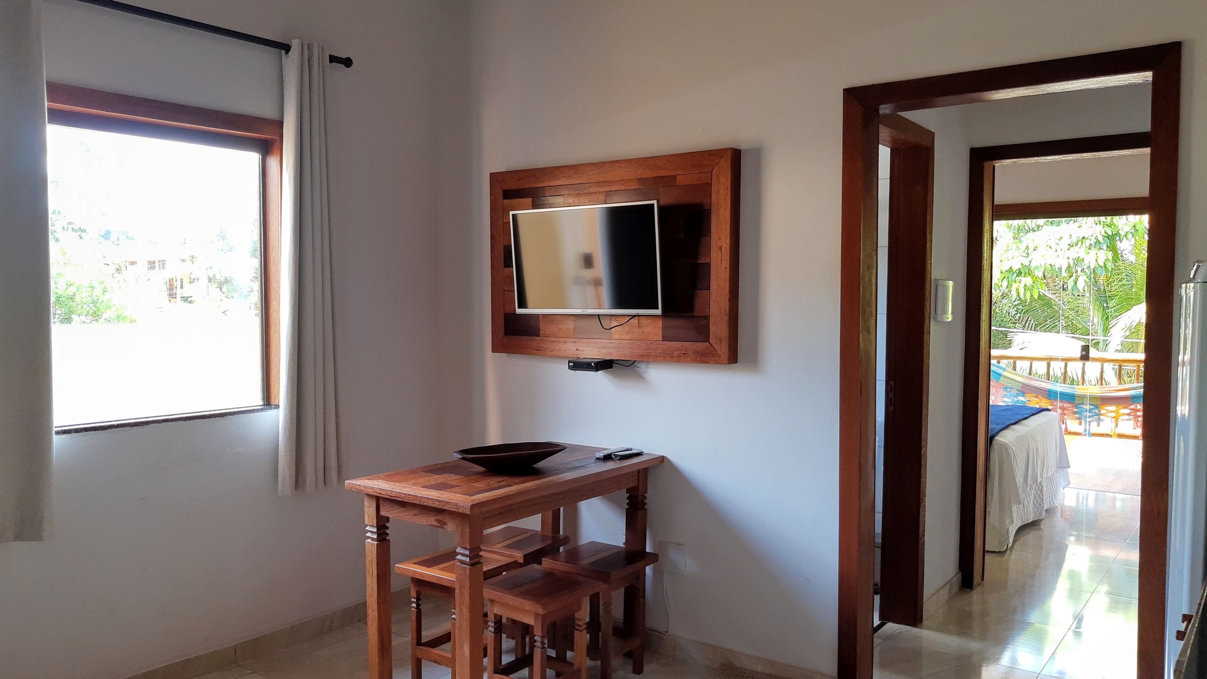 A room with a wooden table and chairs, a television mounted on the wall, and a window with curtains.