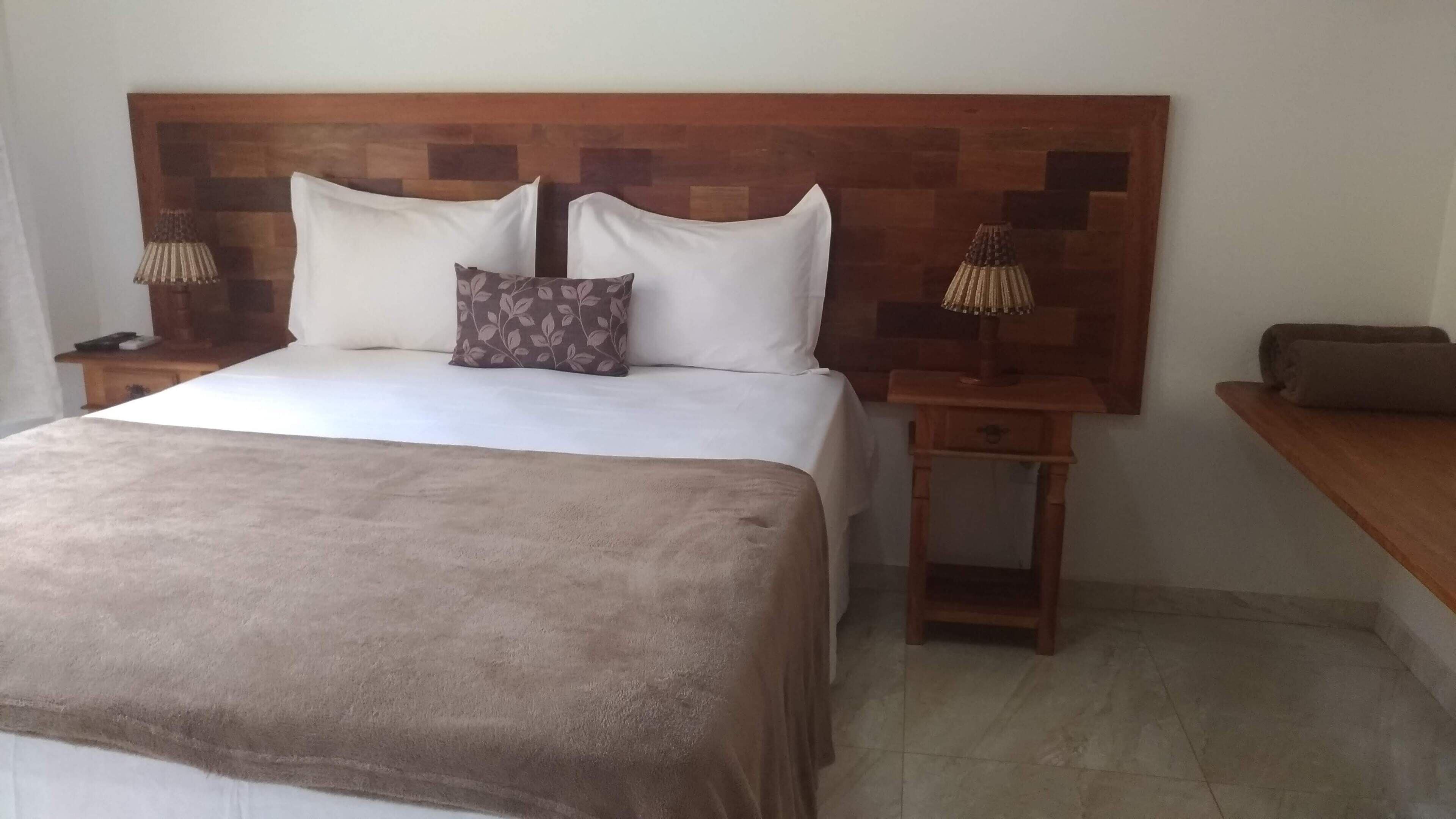 Cama bem arrumada com manta marrom, duas almofadas brancas e uma almofada decorativa com padrão de folha. Há duas mesas de cabeceira de madeira, uma de cada lado da cama.
