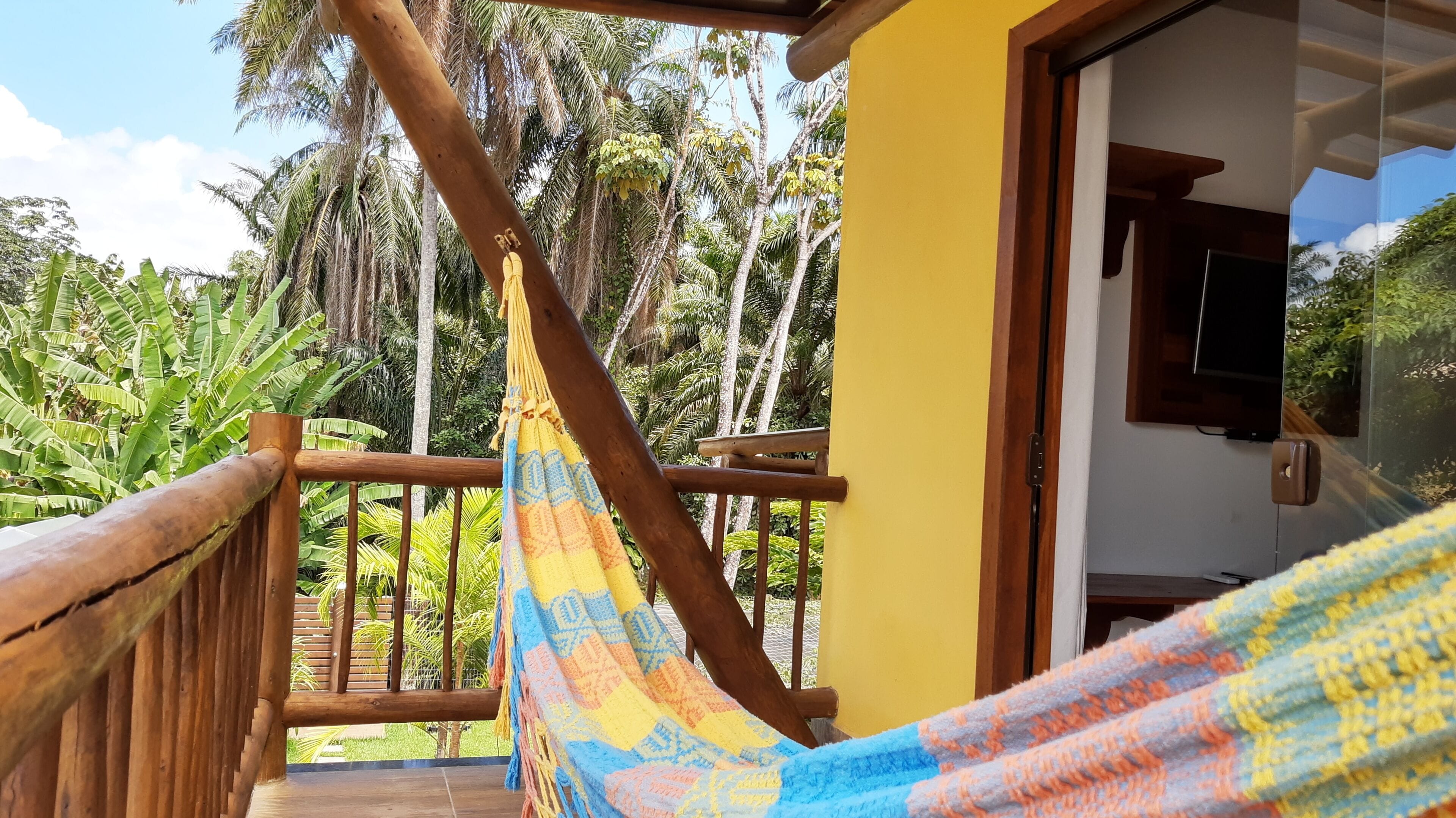 A hammock on a wooden balcony with a view of palm trees and a yellow building.