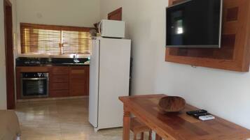 A compact kitchen with a refrigerator, stove, and oven. A wooden table with a bowl and remote controls is in the foreground.