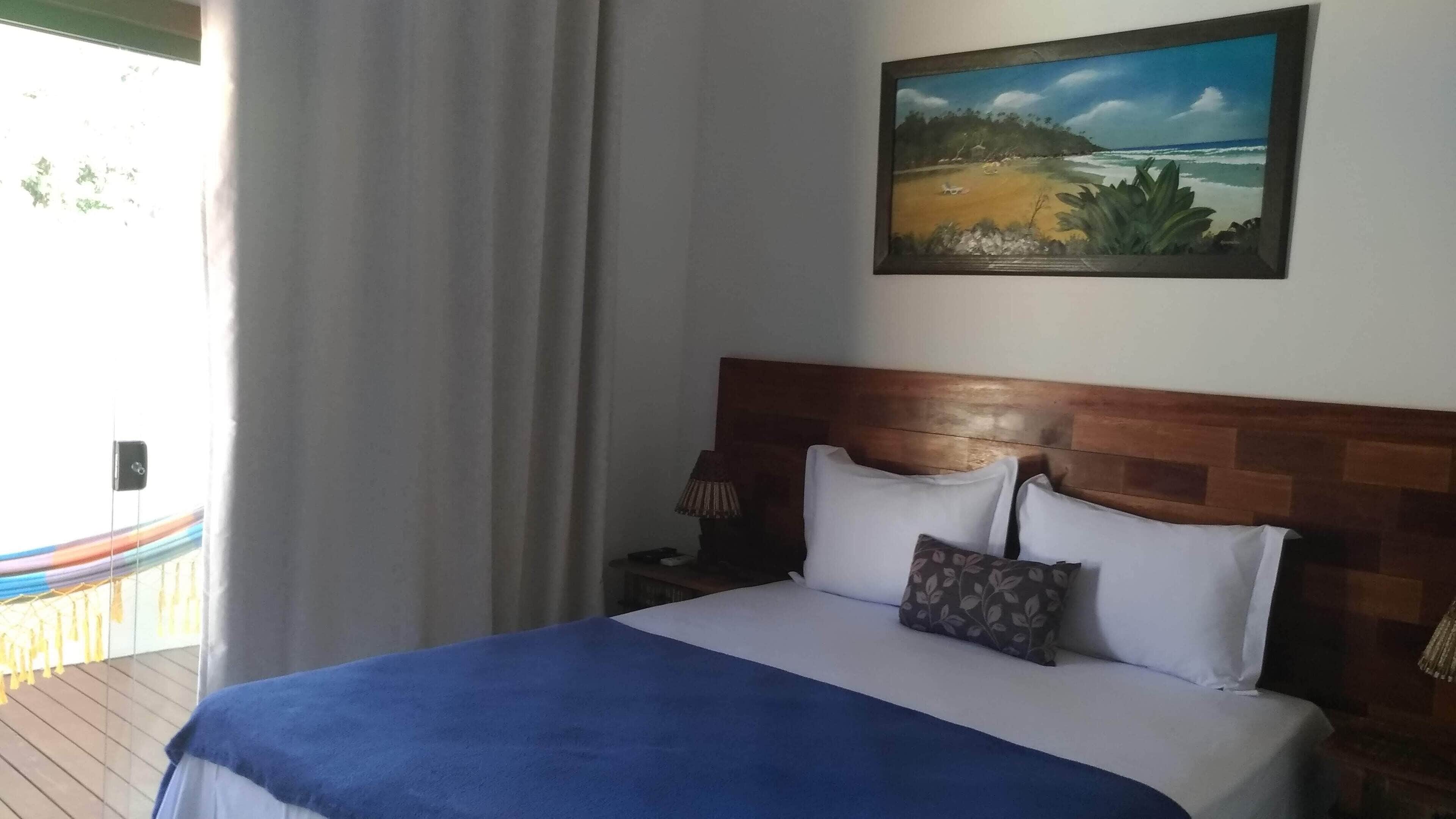 Ein Hotelzimmer mit Bett, einem Strandbild an der Wand, einem hölzernen Kopfteil und einem Fenster mit Vorhängen.