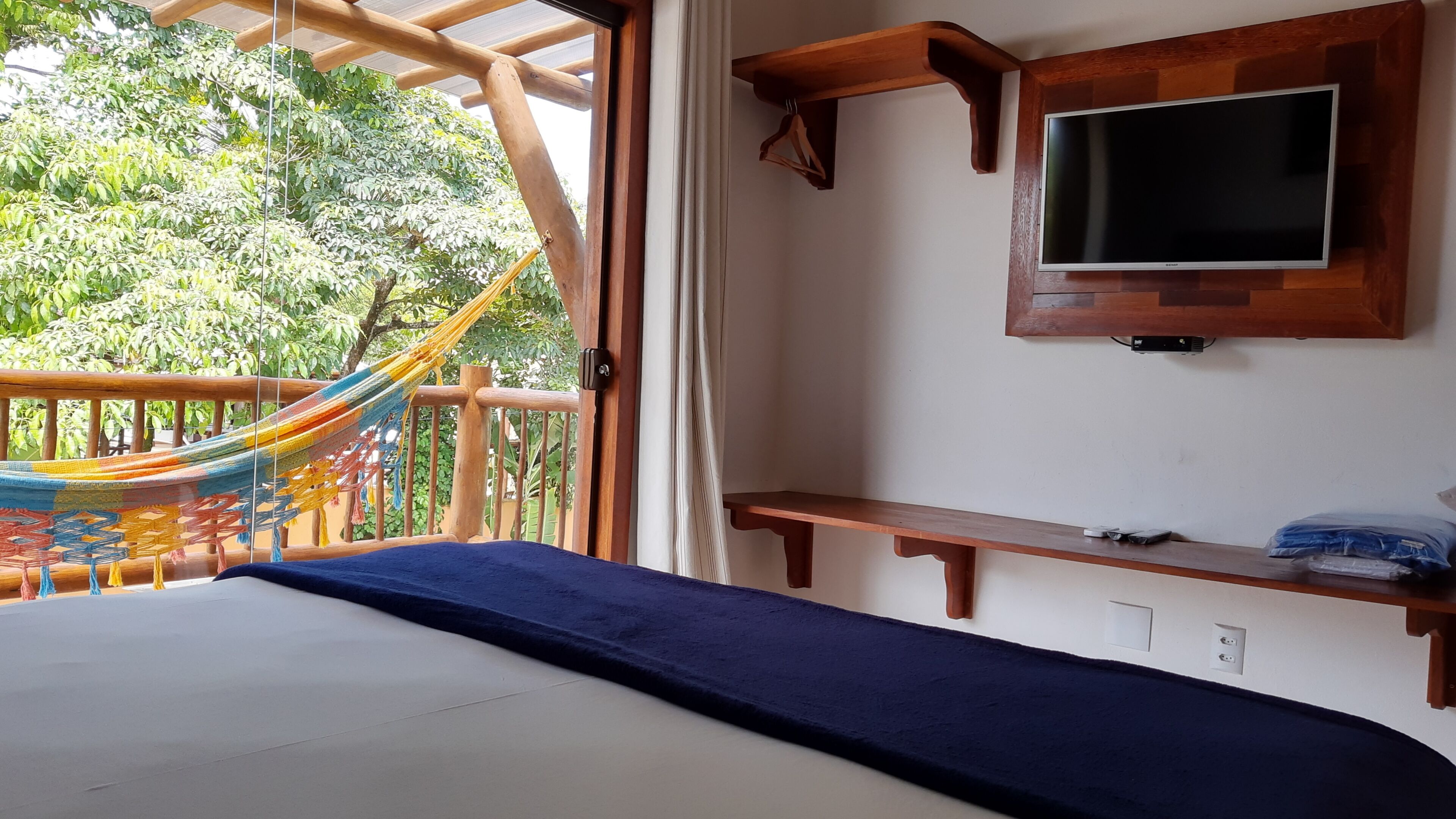 Un dormitorio con cama, televisor montado en la pared, estante de madera y hamaca frente a la ventana.