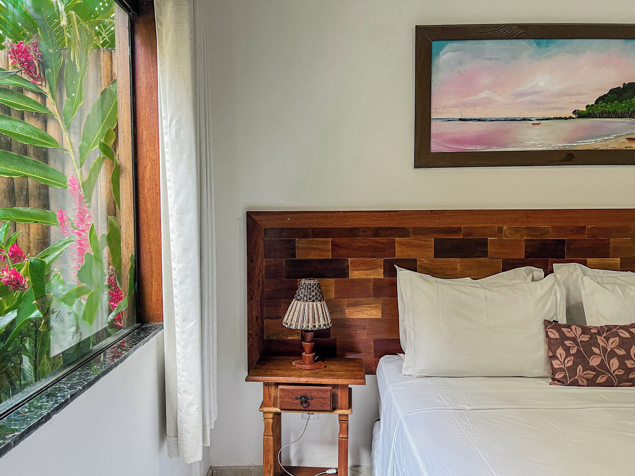 A bedroom with a wooden headboard, a bedside table with a lamp, a bed with pillows, and a window with curtains and a view of greenery.