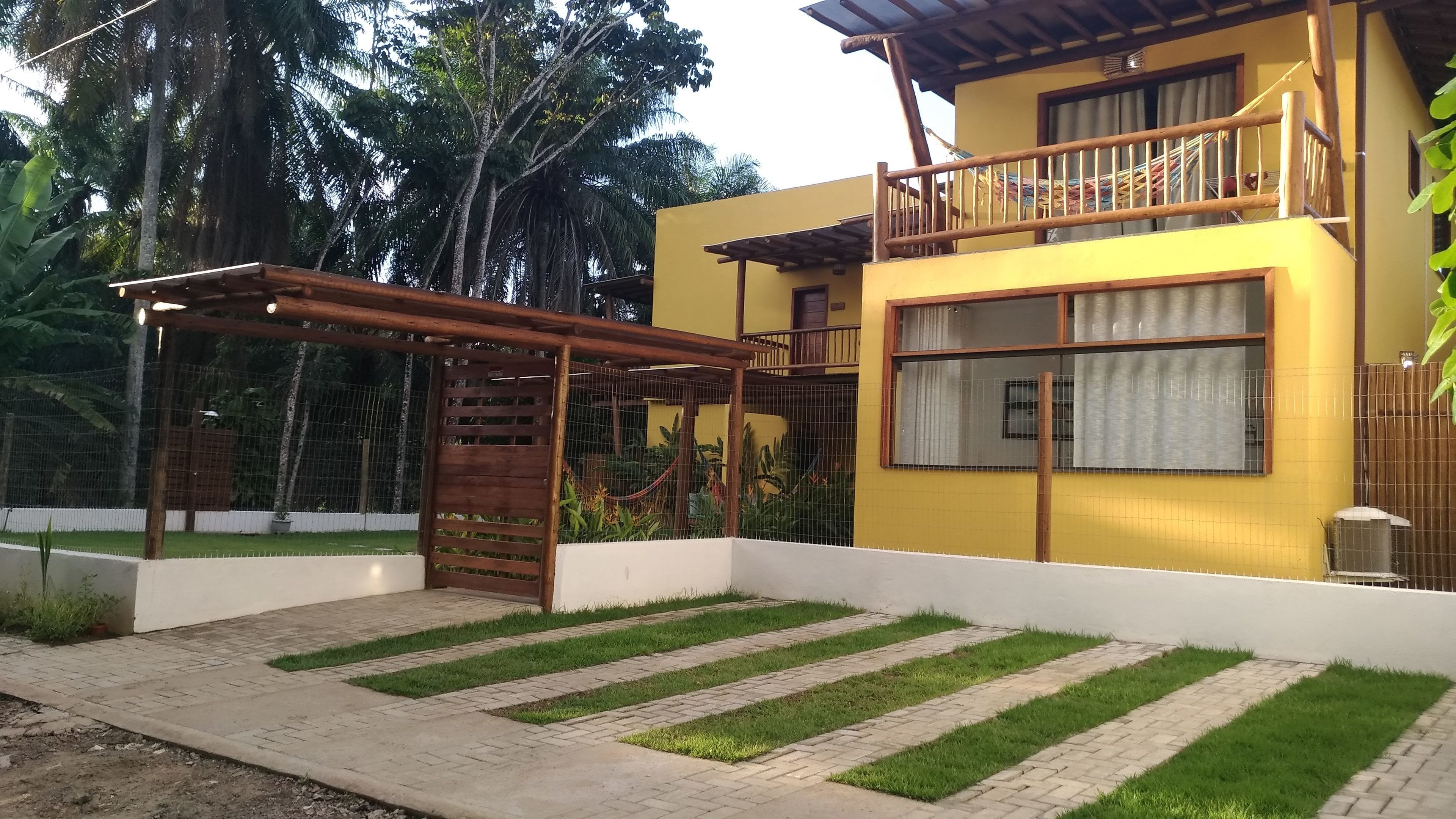 A two-story yellow building with a wooden balcony, a covered patio area, and a fenced garden with grass and plants.