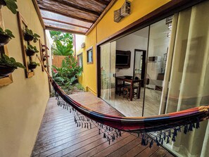 Uma rede em um deck de madeira, uma porta de correr de vidro dando para uma sala de jantar e plantas em vasos na parede.