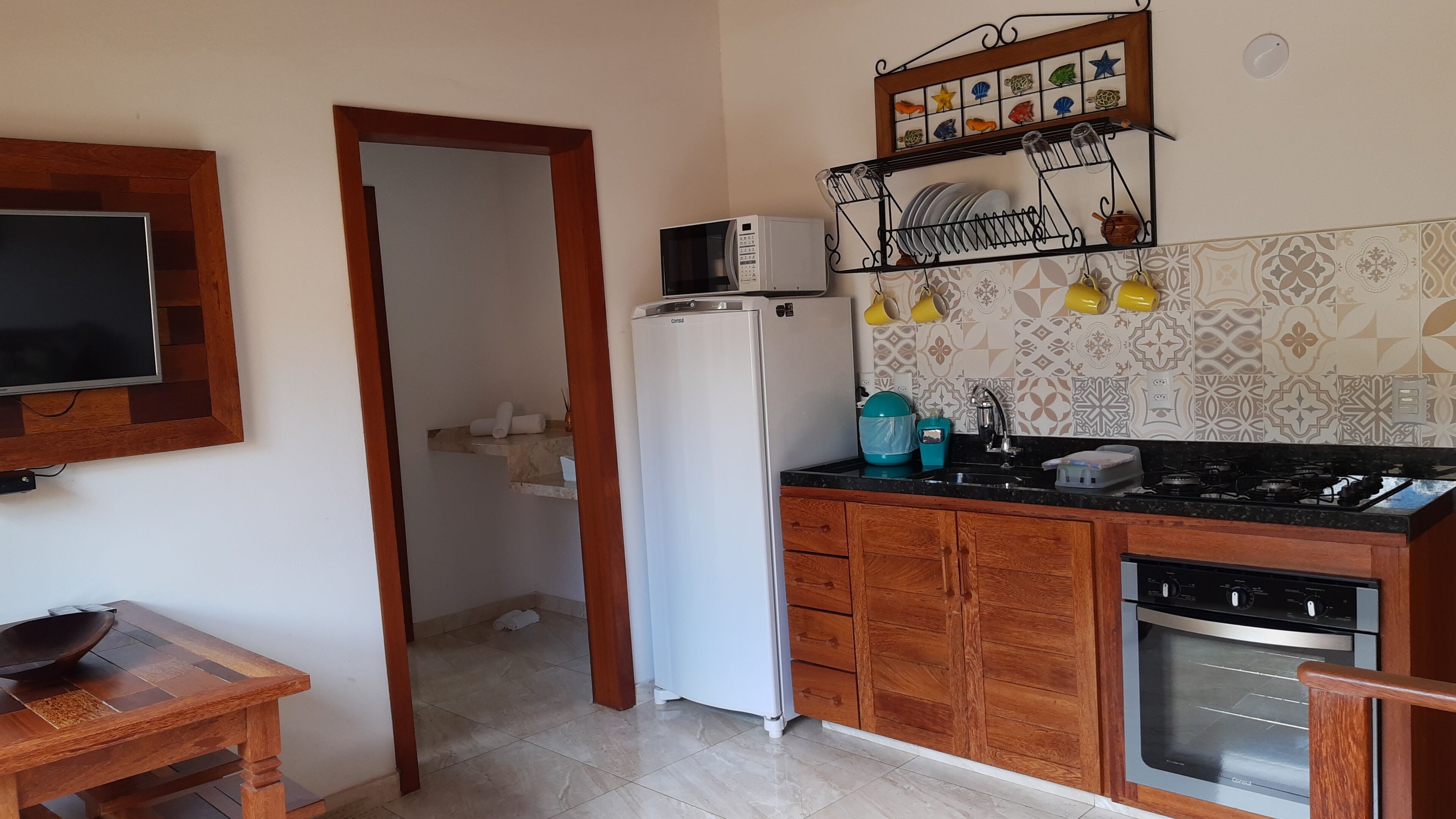 A compact kitchen with a refrigerator, microwave, stove, and sink. There is a wooden table with a bowl and a chair. The kitchen has patterned tiles and a wooden cabinet.