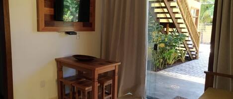 A small wooden table with a bowl, a mounted television, and a sliding glass door leading to an outdoor area with a wooden staircase and plants.