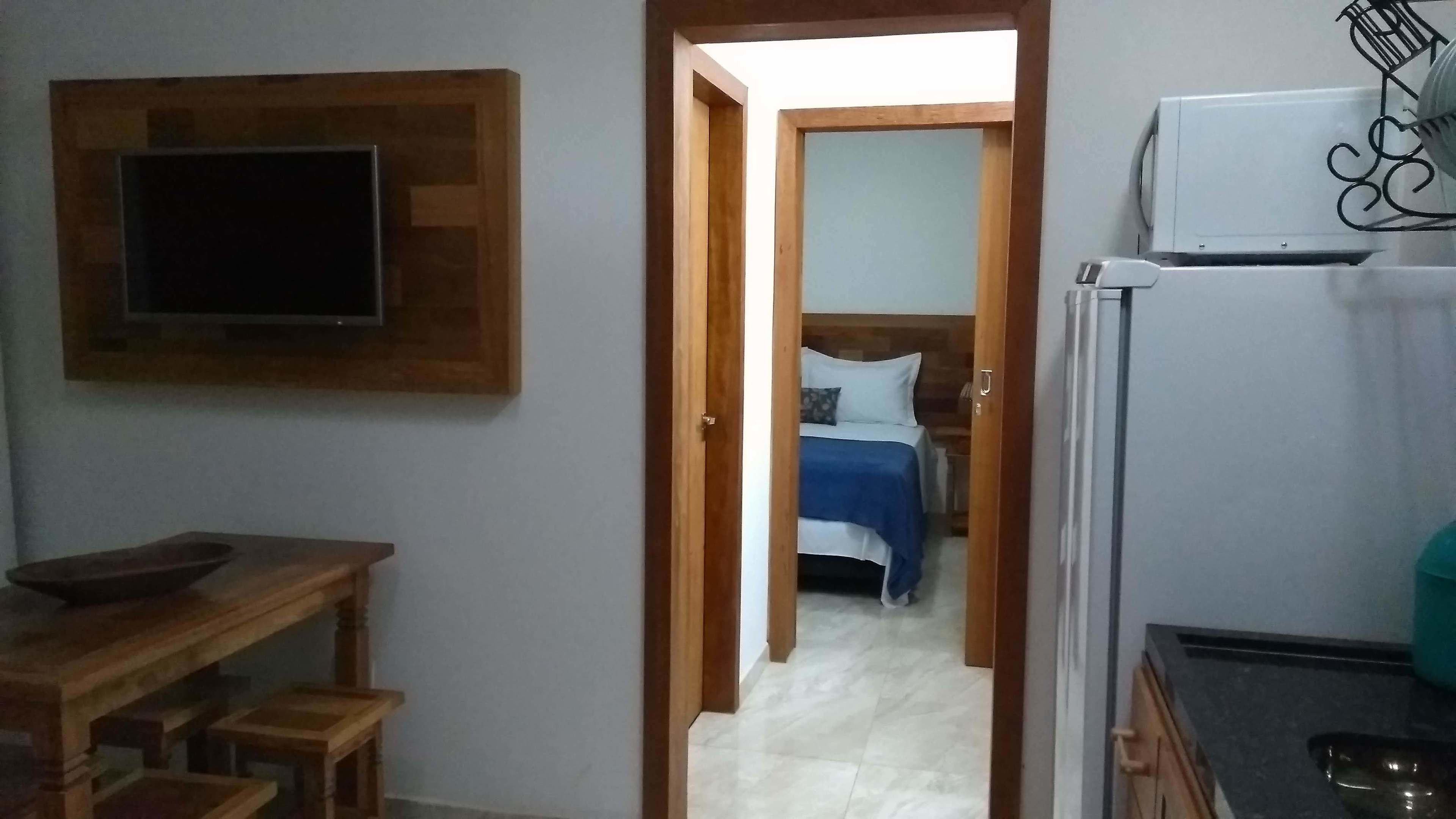 A narrow hallway leading to a bedroom with a bed, a television mounted on the wall, and a refrigerator.