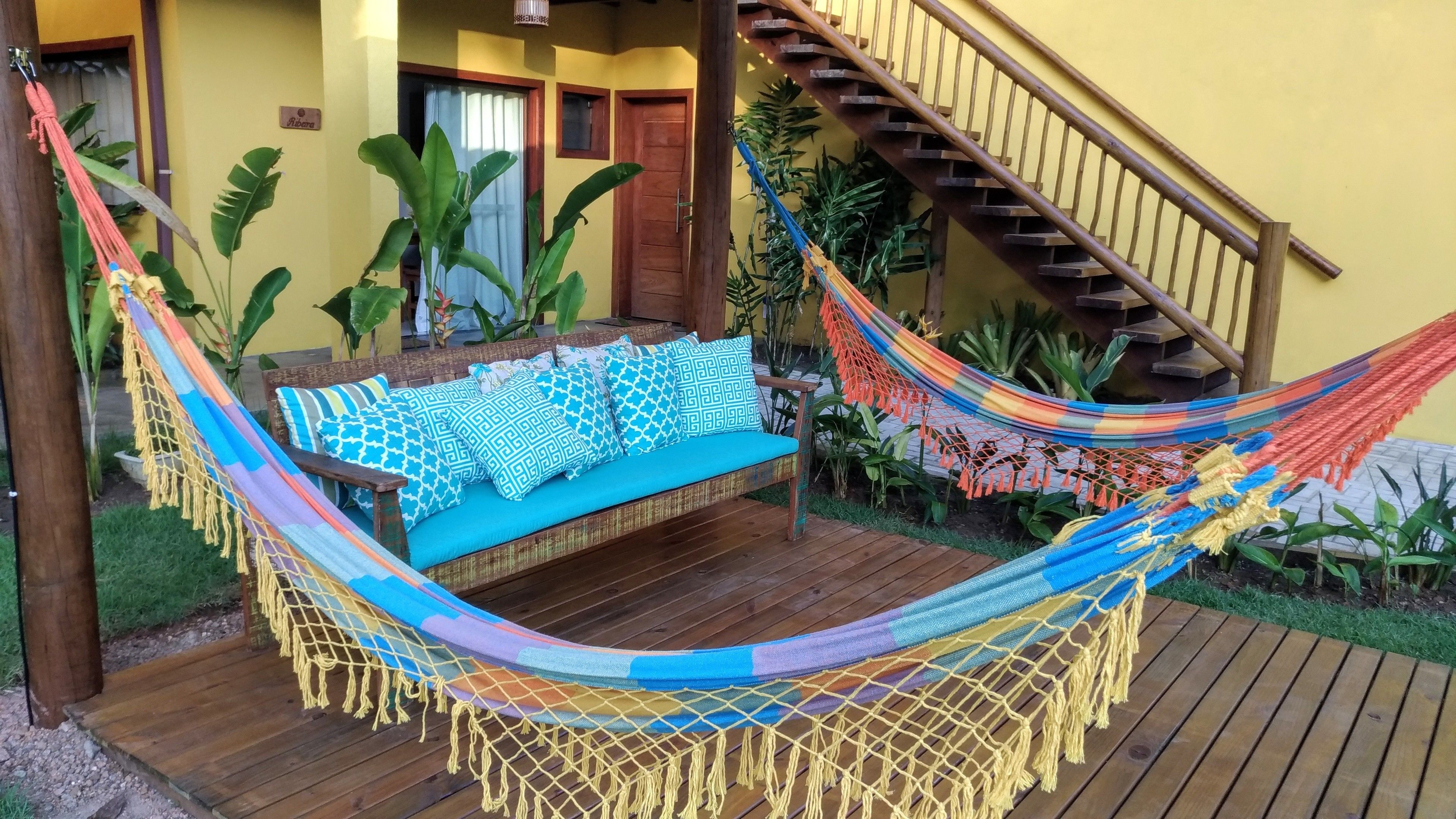 A hammock with a blue cushion and patterned pillows, wooden deck, and a staircase.