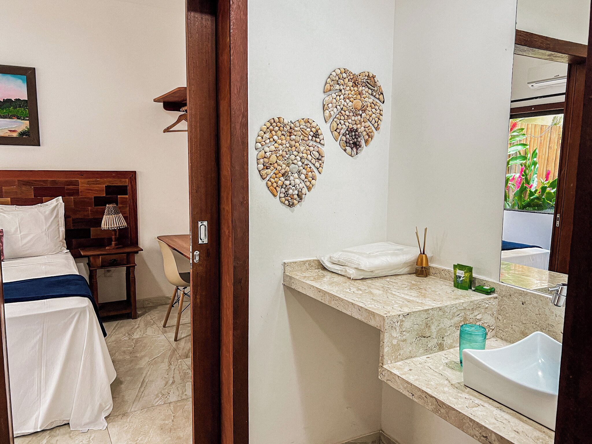 Une chambre d’hôtel comprenant un lit, une table de chevet, un miroir, une salle de bain avec un lavabo et un miroir, ainsi que des coquillages décoratifs accrochés au mur.