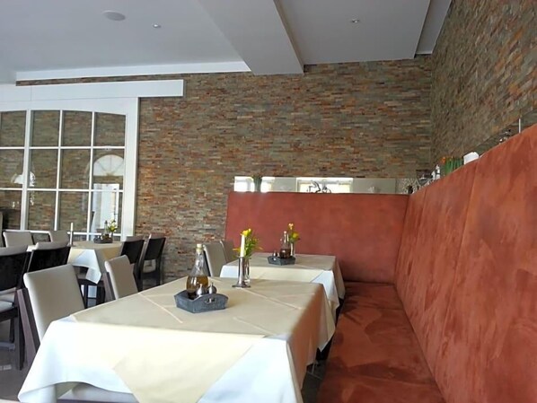 A restaurant with a brick wall, tables with white tablecloths, and a red cushioned bench.