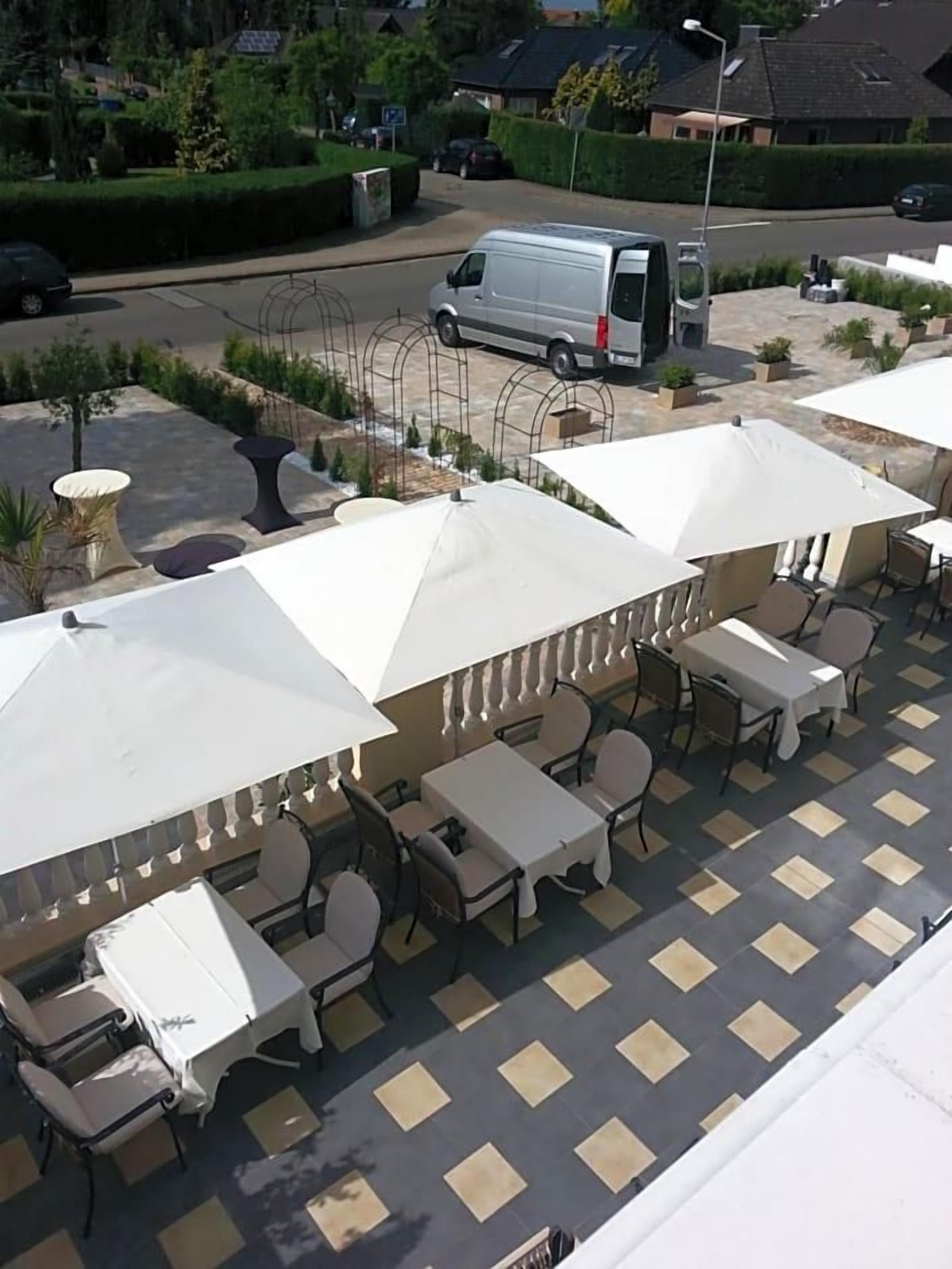 An outdoor seating area with tables and chairs under umbrellas, a parking area with a van, and a hedge-lined street.