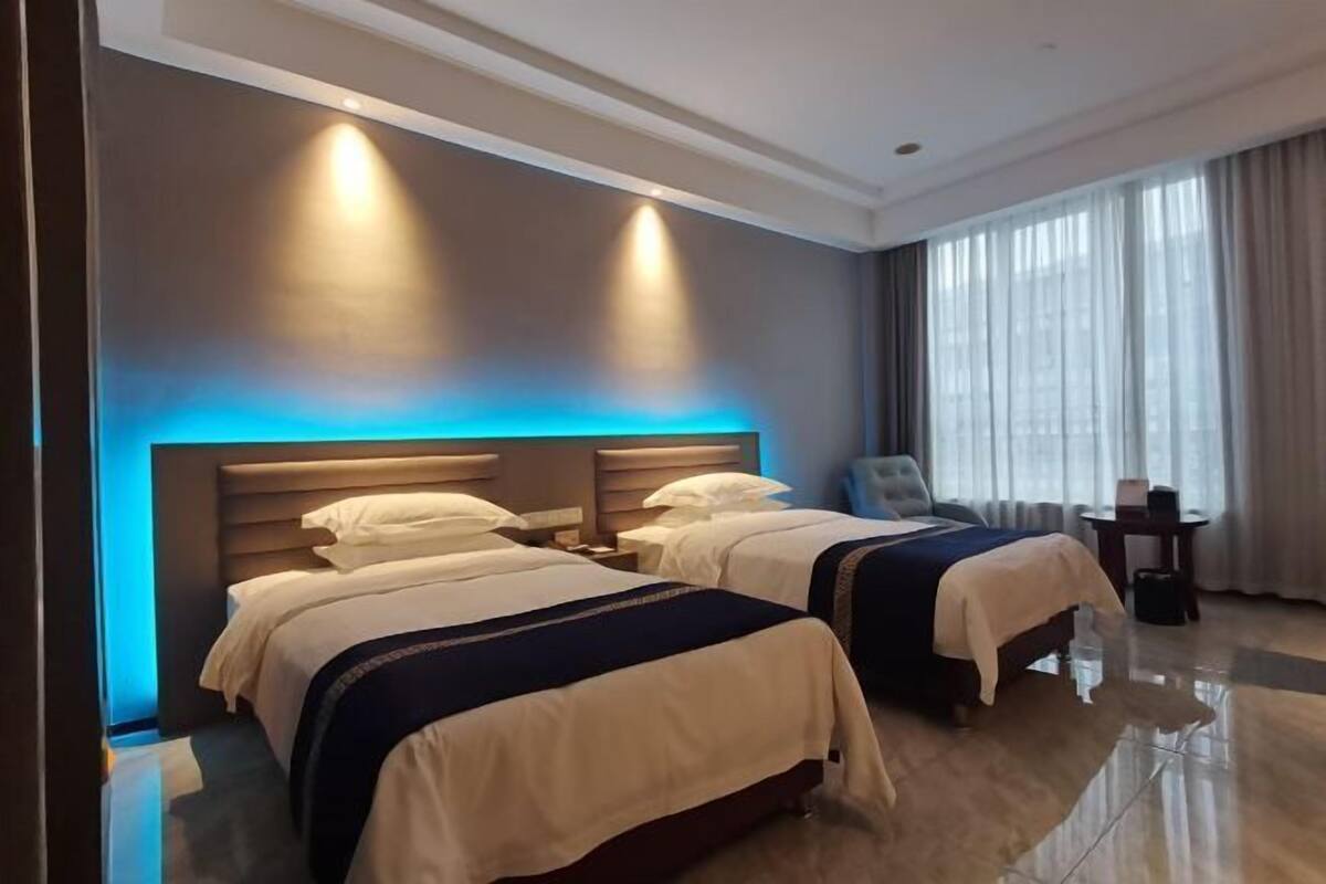 A hotel room with two beds, a chair, a small table, and a large window with curtains.
