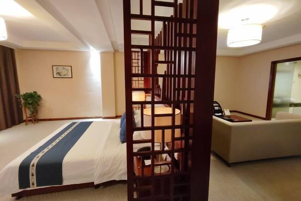 A hotel room with a bed, a desk, a chair, a lamp, and a decorative screen.