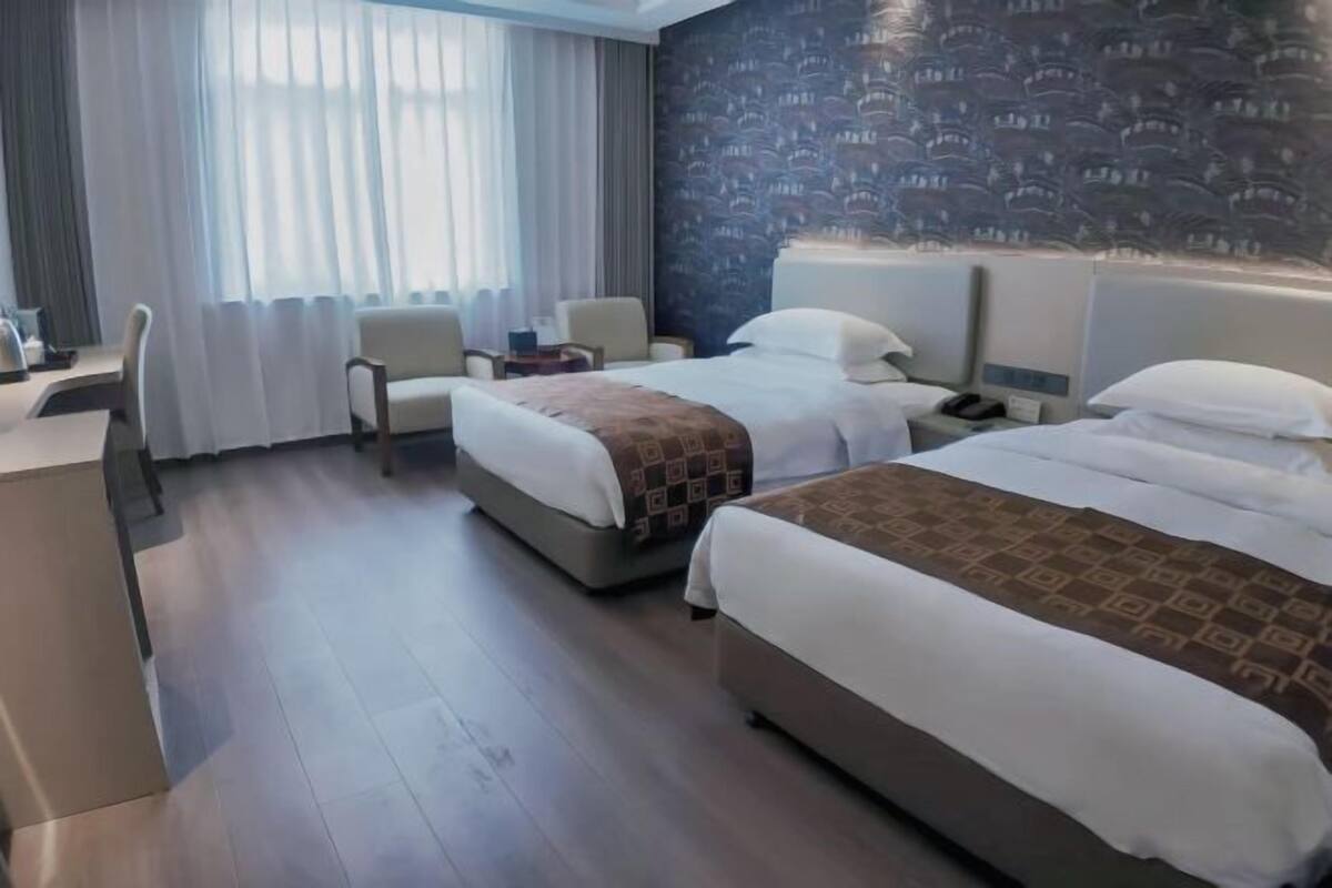 A hotel room with two beds, a desk, chairs, and a large window with curtains.