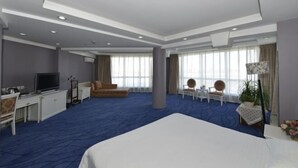Habitación de hotel con una cama grande, un escritorio, un televisor, un sofá y un ventanal con cortinas.