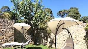 Deux fauteuils de jardin, dotés de parasols, sont placés devant un mur de pierre et entourés de verdure.