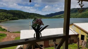 A view from a window overlooking a lake with mountains in the background, a small table with a vase of flowers, and a building with a corrugated metal roof. - Navutovuto Accommodation (Kadavu Island)
