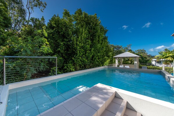 Une piscine dotée d’une terrasse carrelée, entourée d’une végétation luxuriante et surplombée d’un ciel bleu azur.
