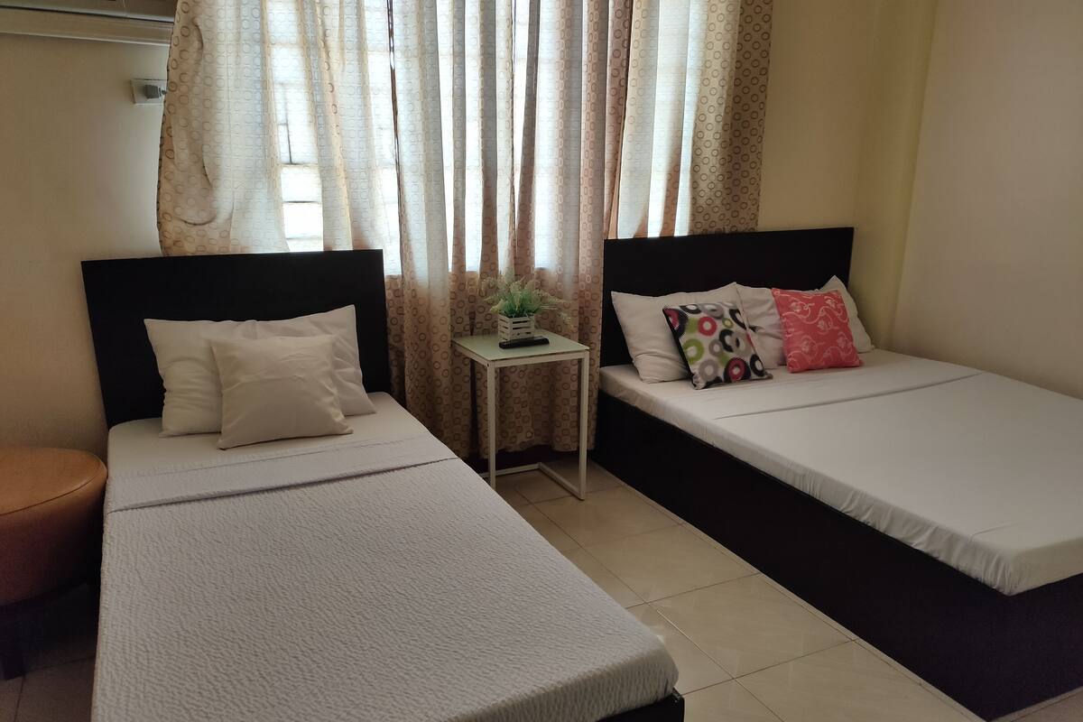 Quarto com duas camas, uma mesinha com uma planta e uma janela com cortinas.