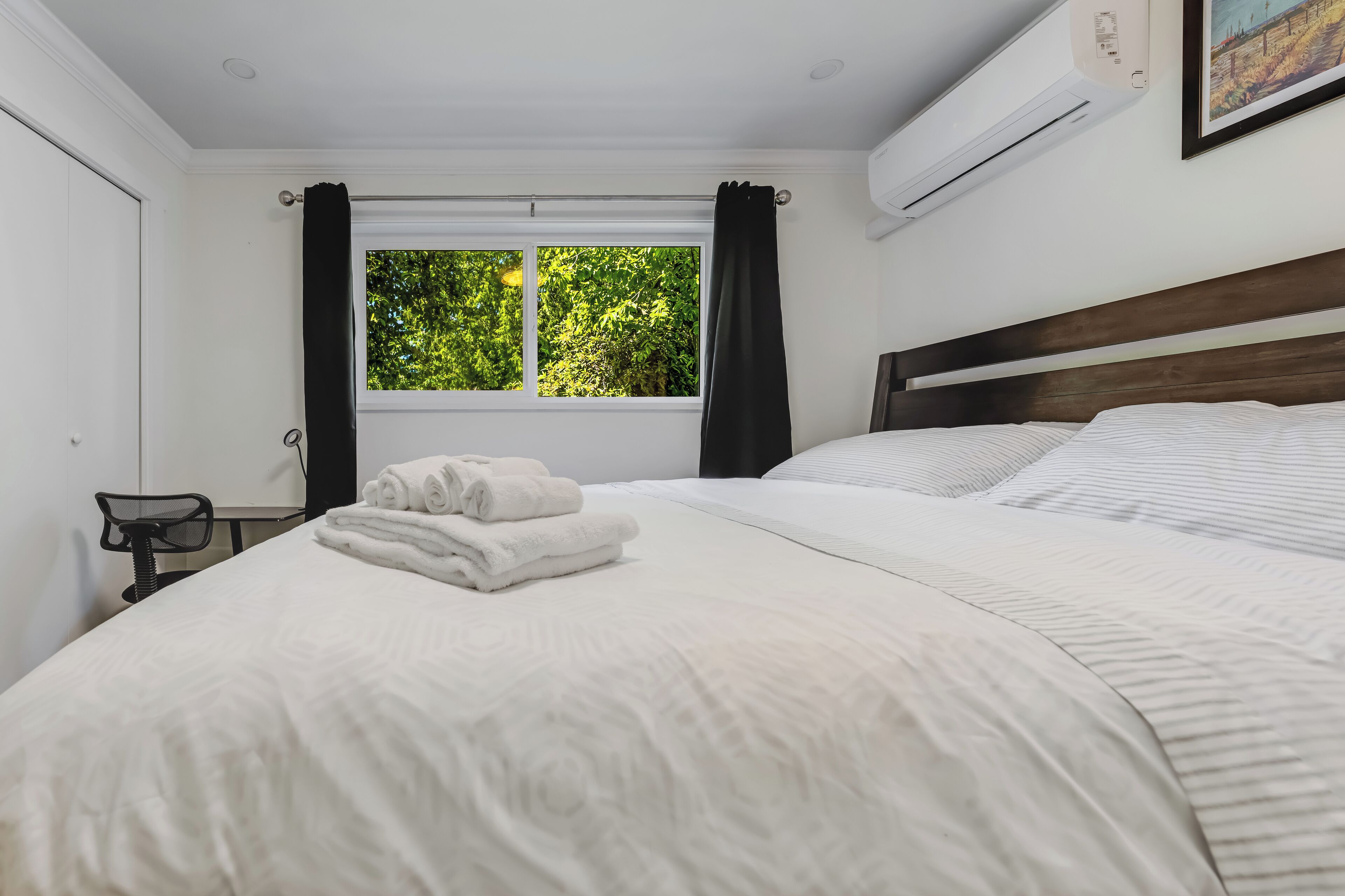 A neatly made bed with white linens and rolled towels, a wooden headboard, a window with curtains, and an air conditioning unit on the wall.