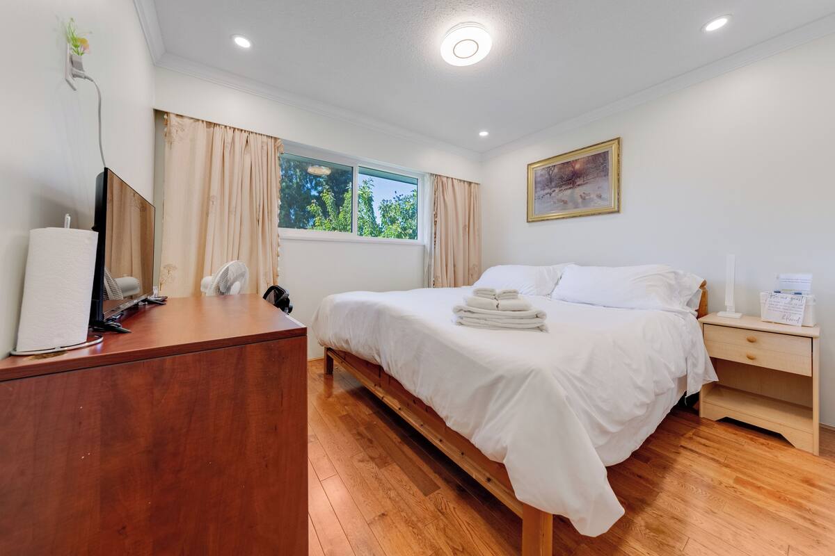 A neatly arranged bedroom with a wooden desk, a bed with white linens, a bedside table, and a window with curtains.