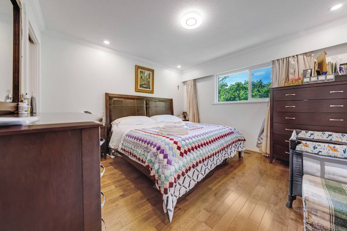 A bedroom with a wooden floor, a bed with a patterned quilt, a dresser, a window with a view, and a nightstand.