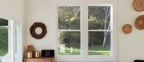 A room with a wooden ceiling, a large window with a view of greenery, two white chairs with cushions, a wooden cabinet, and a wall-mounted air conditioning unit.