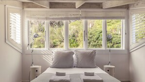 A bedroom with a large window, two bedside tables, a bed with striped pillows, and two towels. - Circular Bay (Waiheke Island)
