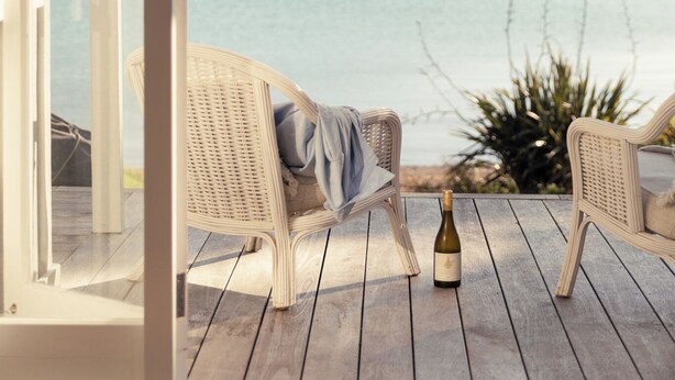 A balcony with wicker chairs, a bottle of wine, and a view of a calm lake and distant hills. - Circular Bay (Waiheke Island)