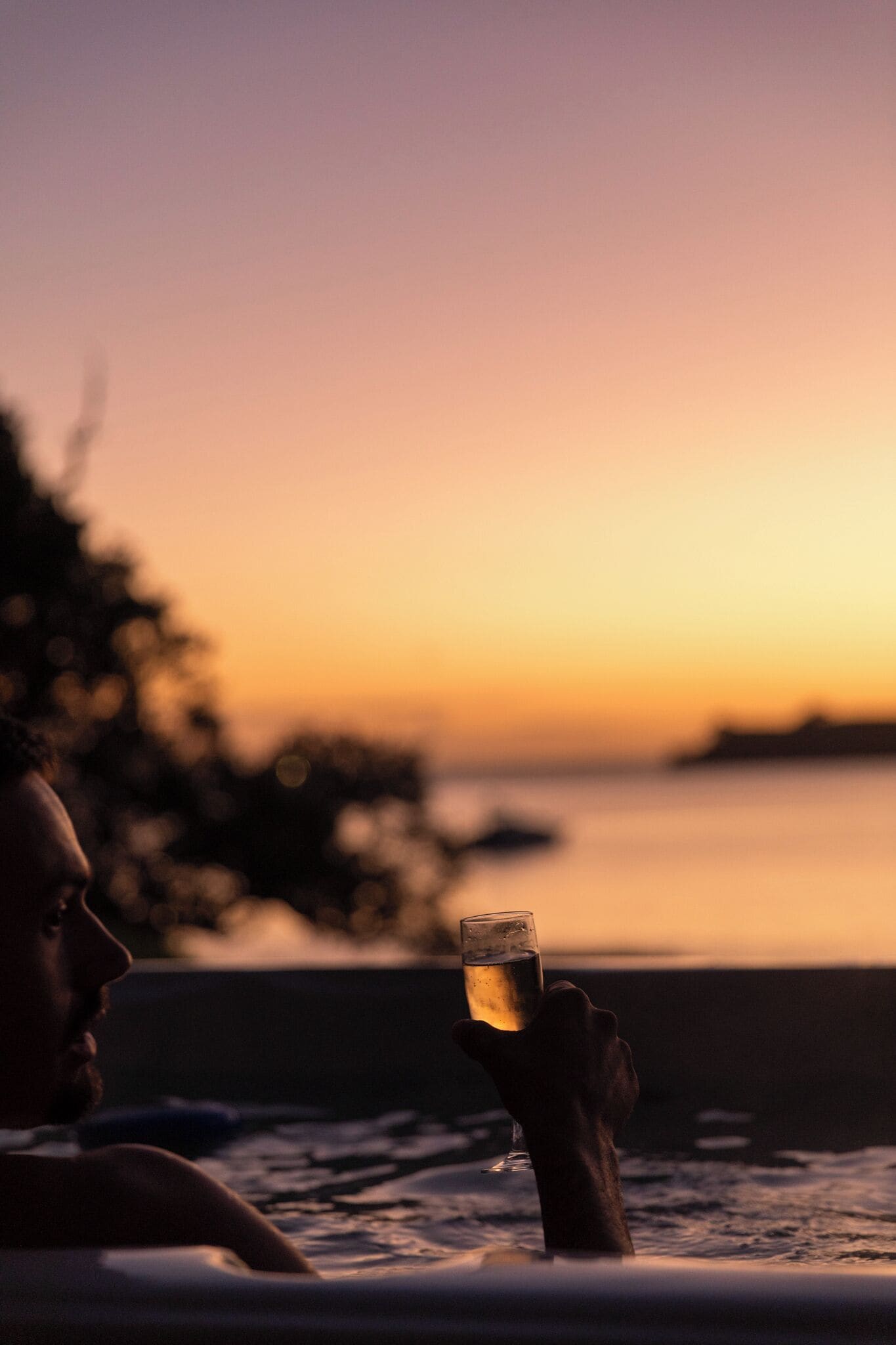 A person holding a glass of drink by a poolside during sunset.