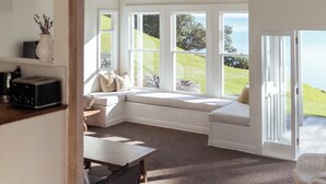 A room with a high ceiling, built-in bench seating, a kitchen area, and large windows offering a view of the outdoors. - Circular Bay (Waiheke Island)