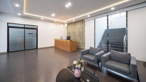 A modern reception area with a reception desk, two grey sofas, a glass coffee table with a vase of flowers, and a staircase in the background. - Moca Hotel (Bangkok)