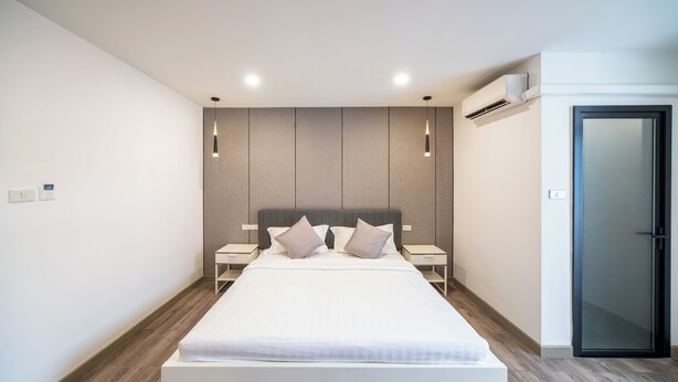 A modern bedroom with a large bed, two bedside tables, a wall-mounted air conditioner, and a door. - Moca Hotel (Bangkok)