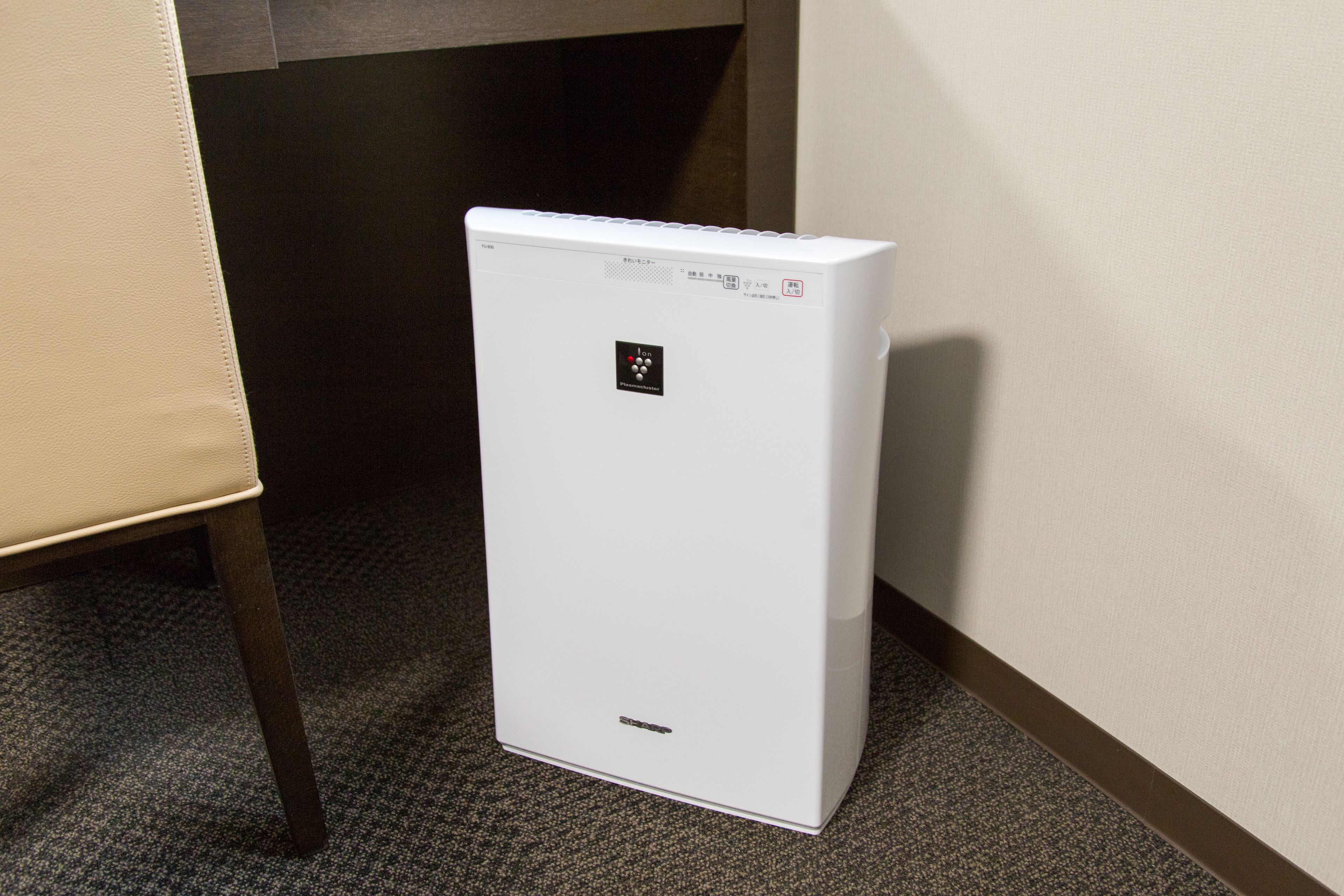 A white portable heater in a corner of a room with a carpeted floor and a chair.