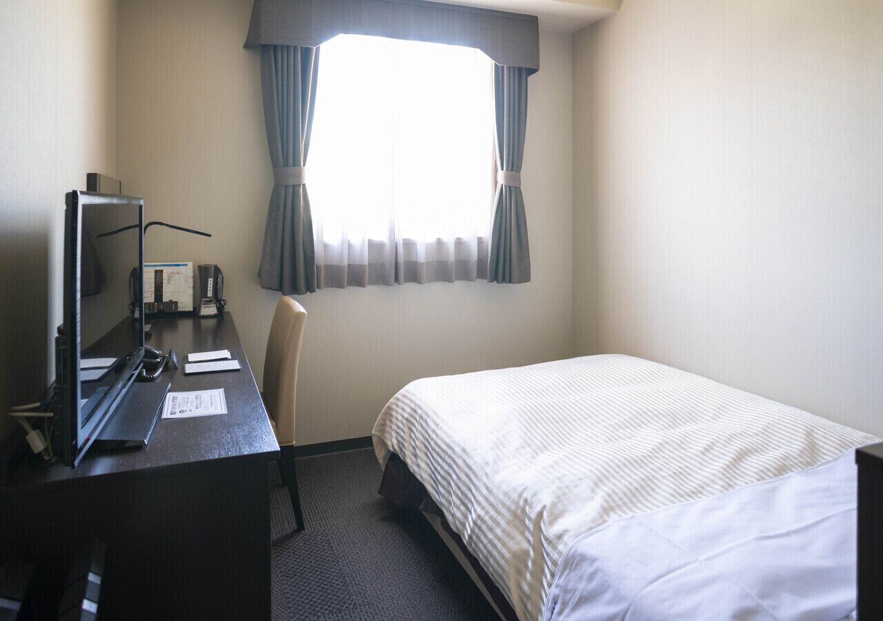 Habitación de hotel con cama, escritorio, computadora, silla y ventana con cortinas.