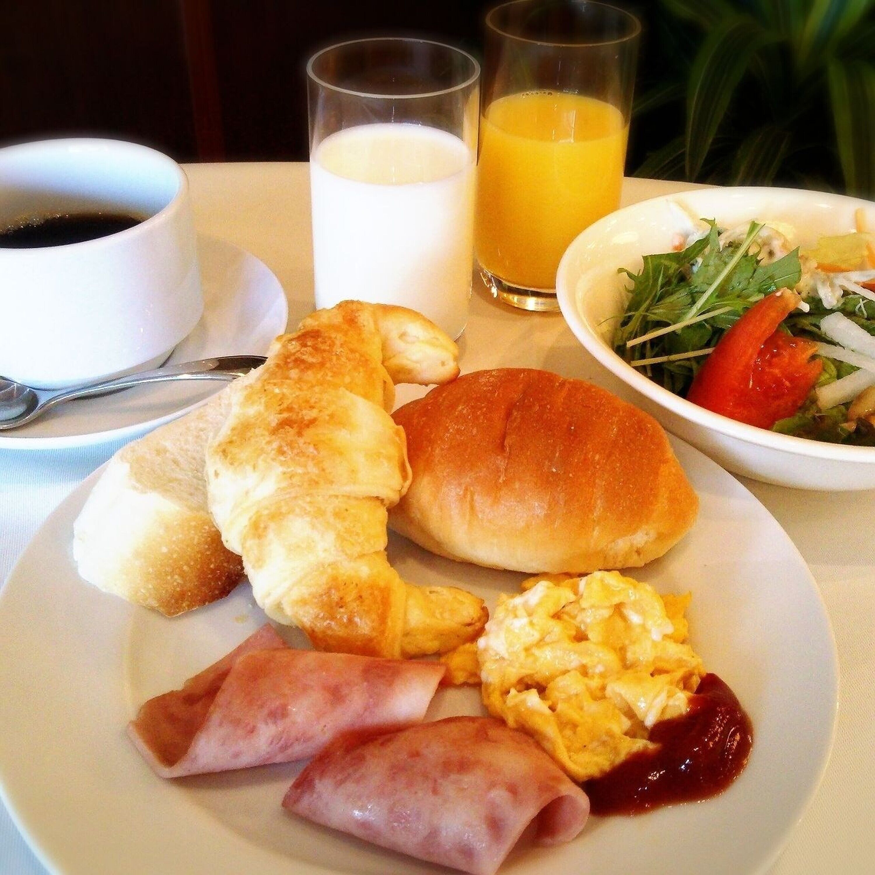 Un plato con cruasanes, un rollo de jamón, huevos revueltos y una ensalada. También hay dos vasos, uno con zumo de naranja y otro con leche.