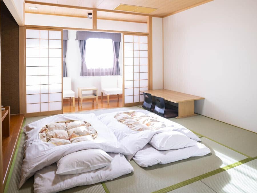 Une pièce de style japonais traditionnel, avec un sol recouvert de tatamis, un futon servant de lit, un petit bureau en bois et un paravent coulissant de type shoji.