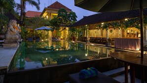 An outdoor swimming pool with a stone statue, a covered seating area, and a building with a tiled roof in the background. - Ubud Aura Retreat (Ubud)