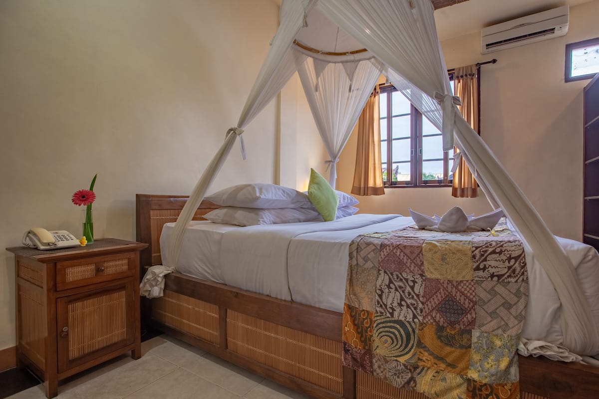 A bedroom with a wooden bed, white bedding, a canopy, a nightstand with a phone, and a window with curtains.