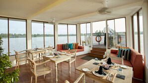 A spacious living area with a dining table, sofas, and large windows overlooking a body of water. - Flow by The Amber Collection - Luxury River Cruises in Sri Lanka (Piliyandala)