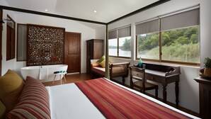 A bedroom with a large bed, a desk, a chair, a window with a view of greenery, and a bathtub. - Flow by The Amber Collection - Luxury River Cruises in Sri Lanka (Piliyandala)