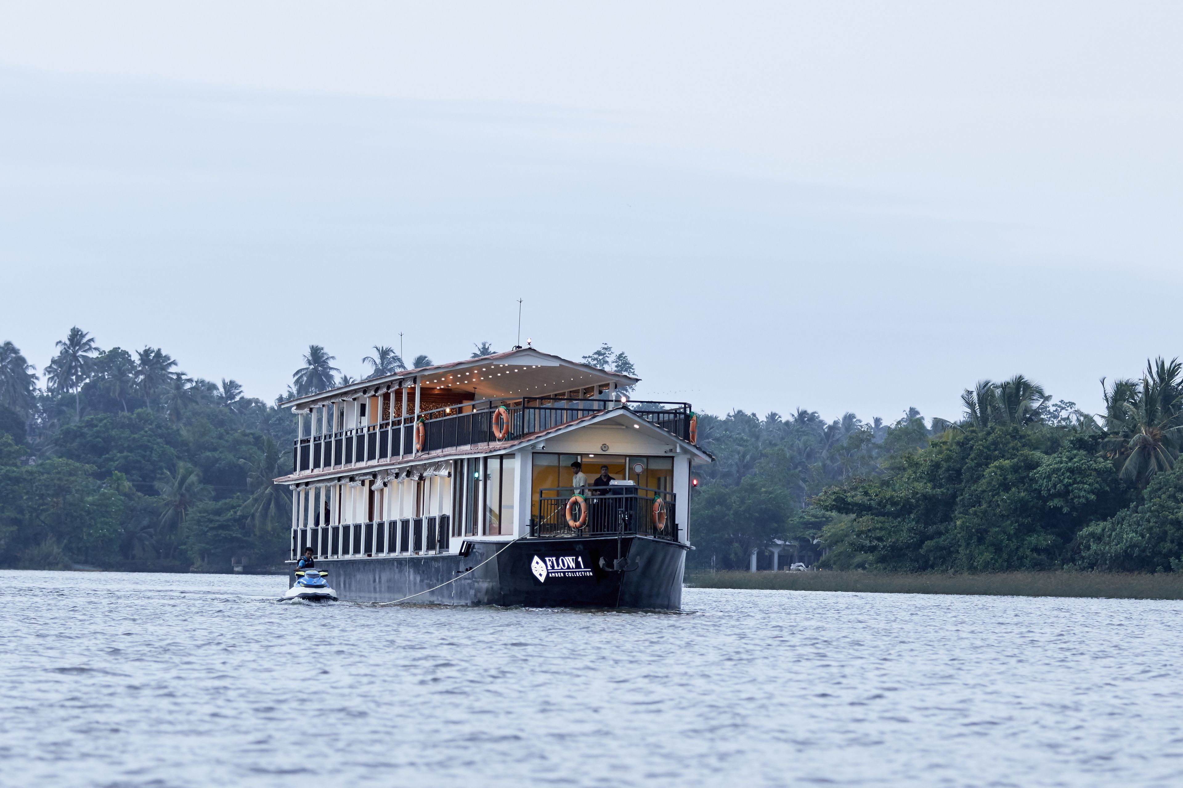 A large houseboat with a balcony and lifebuoy, floating on water with a backdrop of palm trees.